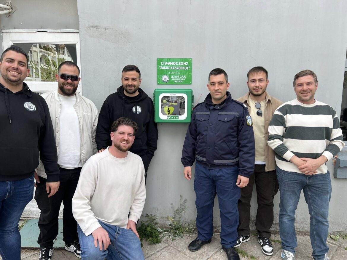 Eight men pose outdoors beside a green AED cabinet mounted on a gray wall, smiling at the camera.
