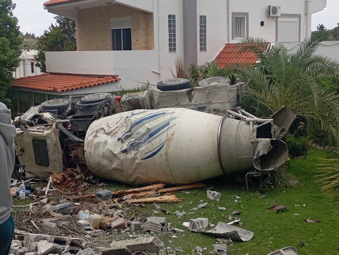 Overturned white cement mixer truck with blue stripes, crashed in a yard with debris and damaged surroundings