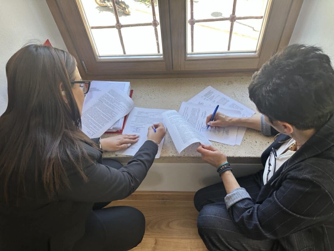 Two people reviewing documents at a windowsill, each writing with a pen and papers spread between them.