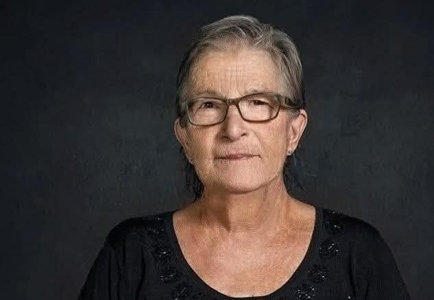 Portrait of an older woman with short gray hair and glasses, wearing a black top, facing the camera against a dark backdrop, with a calm expression.