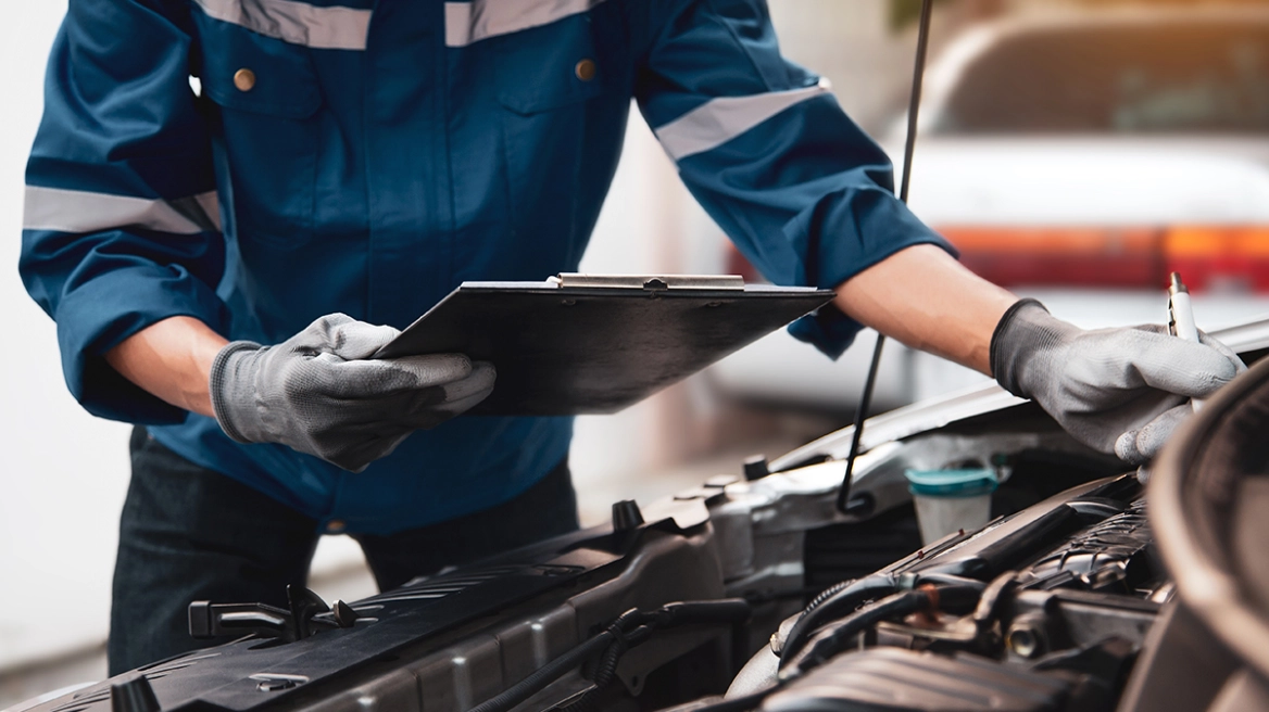 Auto mechanic in a blue uniform and gloves holds a clipboard while inspecting a car engine under the open hood.