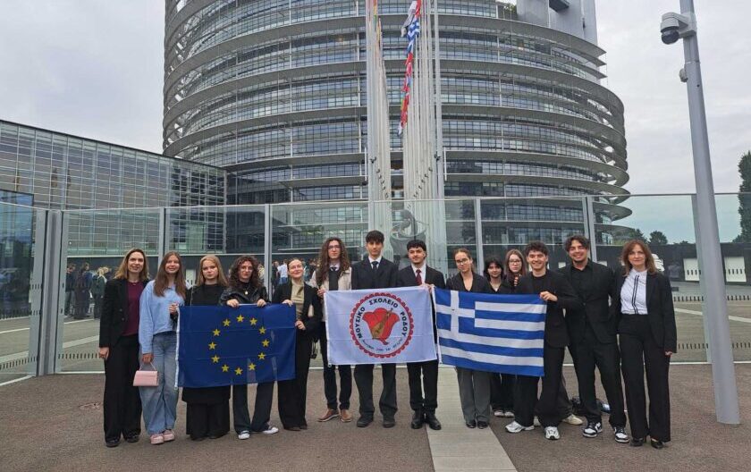 Group of students posing outdoors in front of a curved glass government building, holding the European Union flag, a white banner with a red emblem, and the Greek flag.