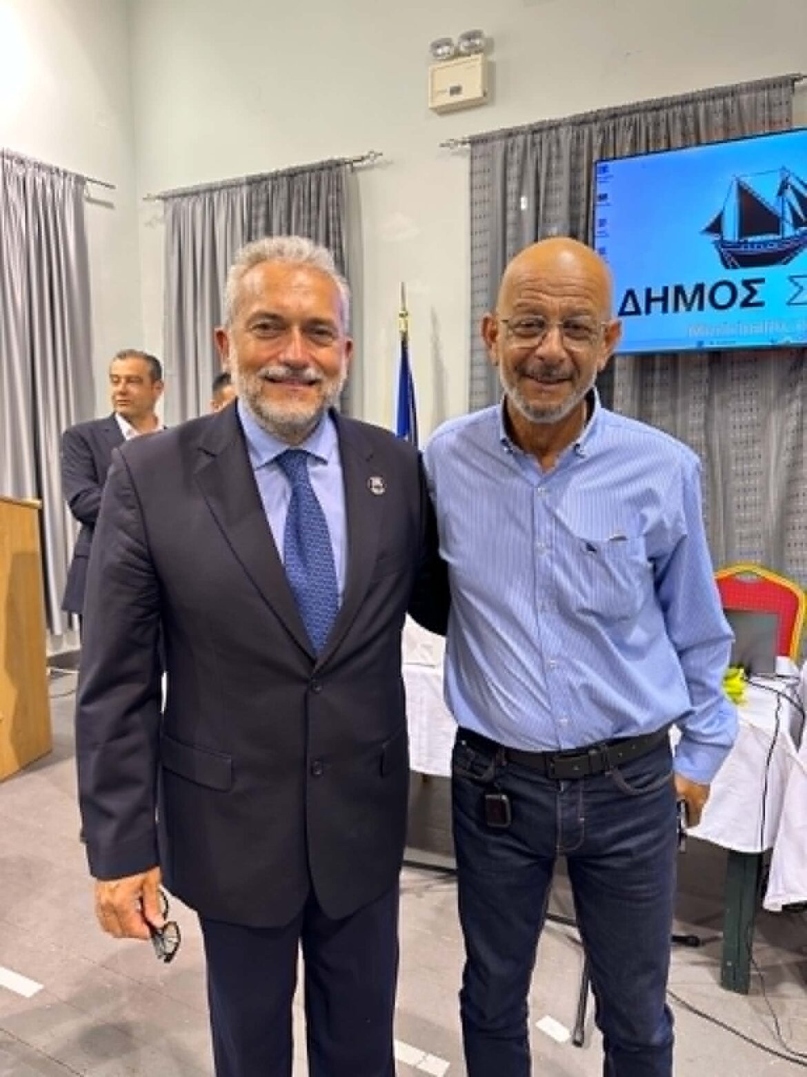 Two smiling men posing indoors, one in a dark suit with blue tie and the other in a light blue shirt, standing side by side in a conference room.