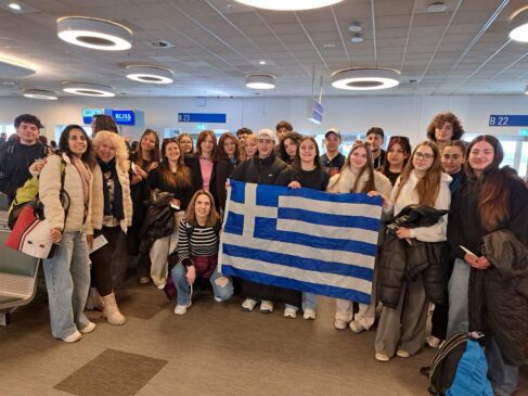 Group of travelers posing in an airport terminal, holding a blue-and-white Greek flag.