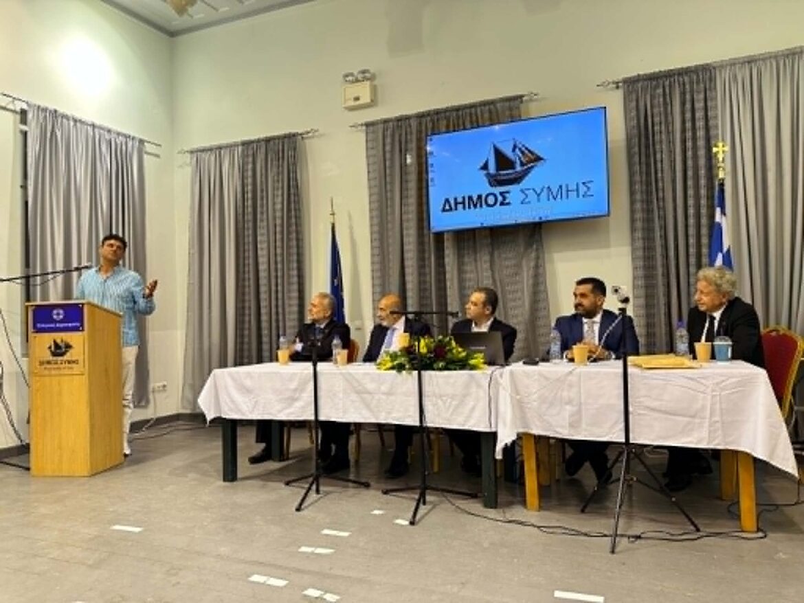 Speaker at podium addressing a panel of officials seated at a long table in a conference room while a screen displays Greek text.