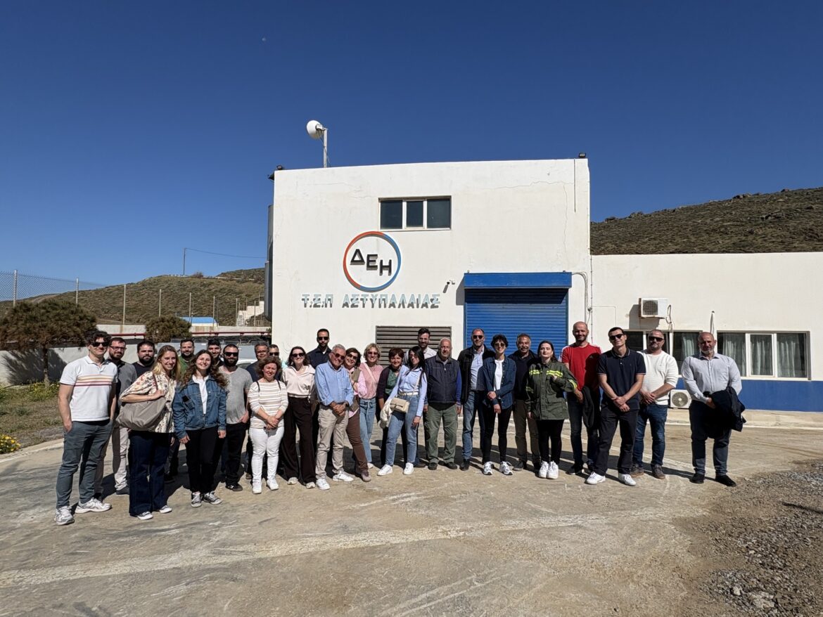 Group of about 25 adults posing outside a white building with a blue roll-up door and Greek lettering on the wall, under a clear blue sky.