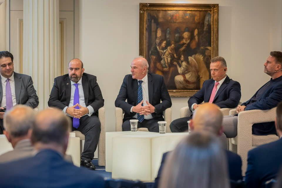 Five men in suits sit on a panel in a formal meeting room, with a large painting behind them and an audience in the foreground.