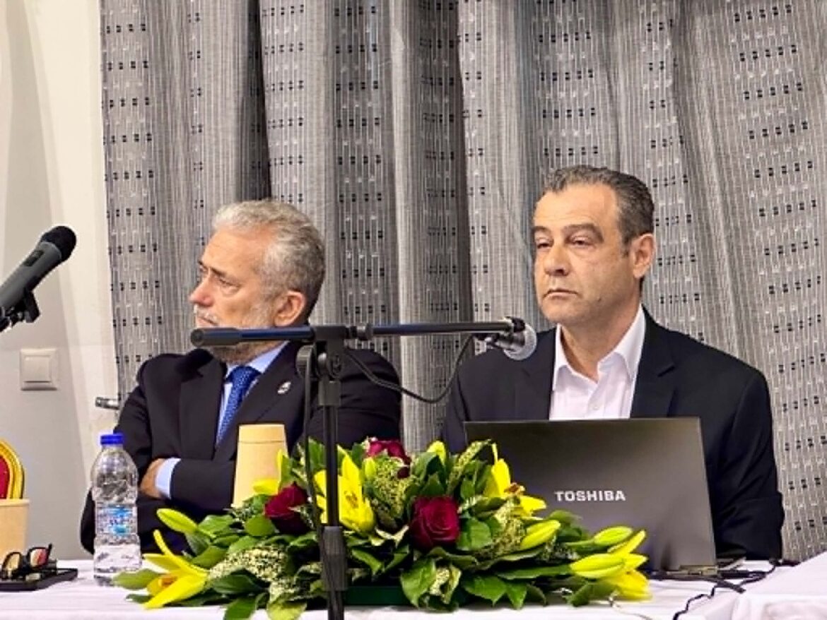 Two men in suits seated at a panel with microphones, a Toshiba laptop, and a colorful flower arrangement in front of them.