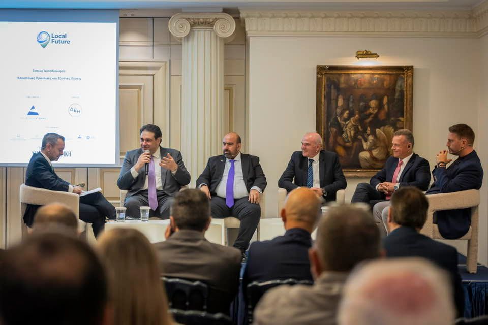 Five men in suits sit on a panel stage in a formal room, with an audience watching; a screen on the left displays the Local Future logo and sponsor logos behind them.