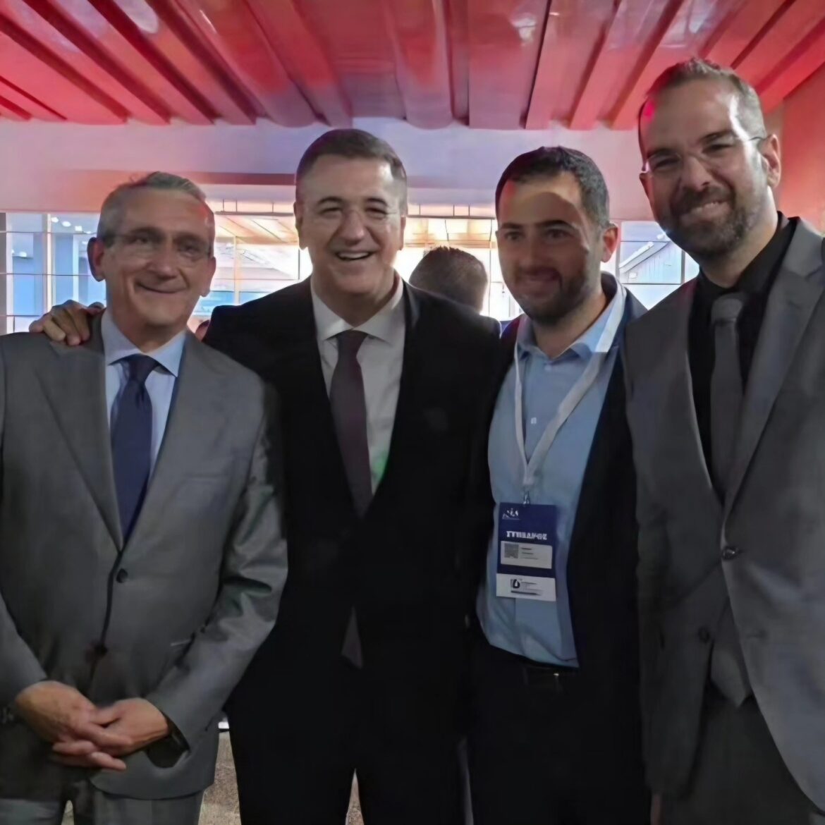 Four men in suits posing together at a formal event, smiling for the camera in an indoor venue with red lighting above them., Four men in business attire at a conference, one wearing a lanyard badge, posing for a group photo., Portrait of four male colleagues in suits standing close together for a photo at a professional event., Four professionals in formal wear posing indoors during a networking or corporate gathering., Smiling group photo of four men in suits at a corporate event, standing side by side.