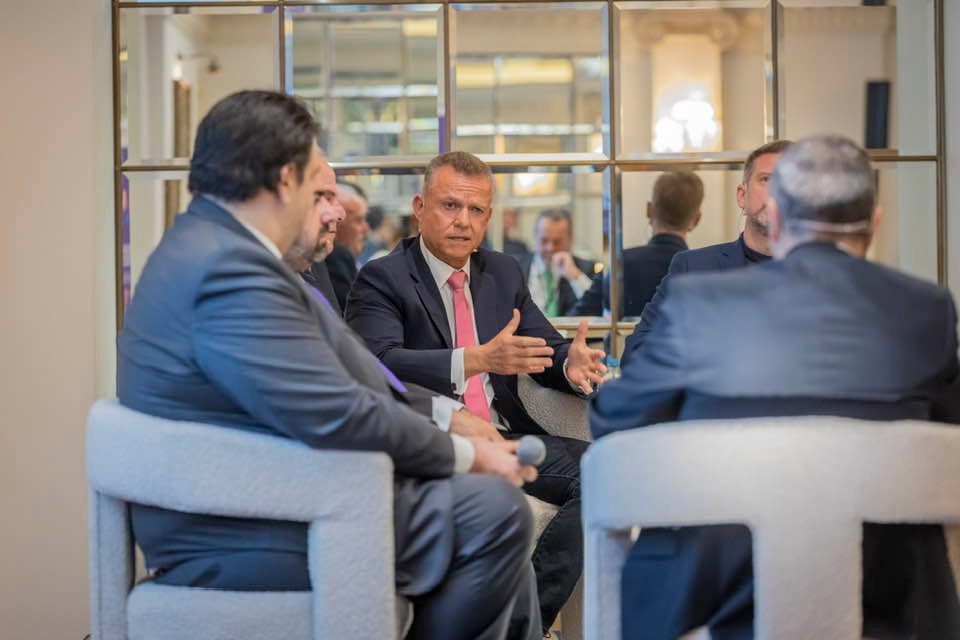 Group of suited men seated in a panel-style discussion; the central man speaks with animated hand gestures, others listening.