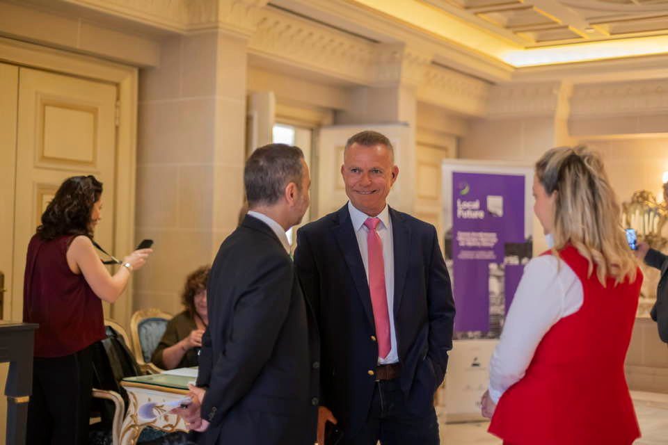 Three professionally dressed adults chat in a bright hotel lobby; man in a navy blazer and pink tie smiles while another man in a dark suit talks nearby, with a woman in a red vest facing them and a purple event poster in the background.