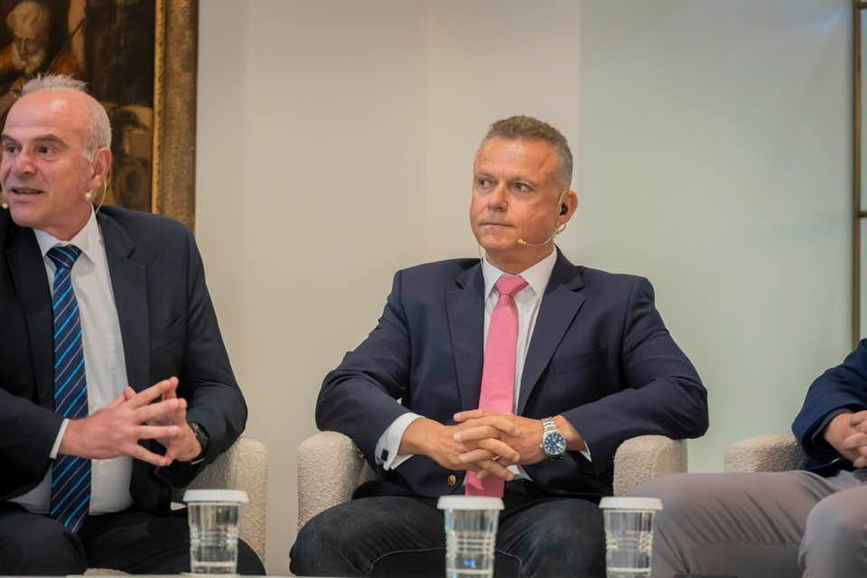 Three men in suits sit on a panel, with two holding hands in lap, microphones on their heads, during a discussion in a formal setting.