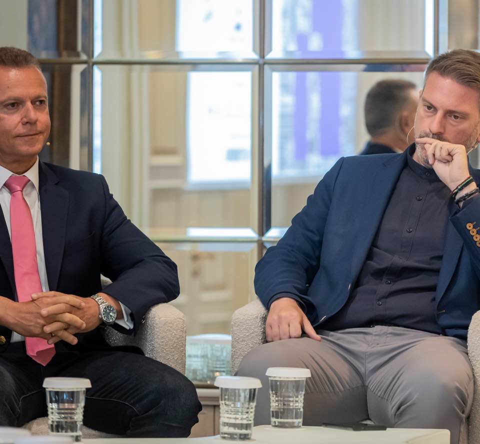 Two men in business attire sit on armchairs in a modern office set, engaged in a discussion, with coffee cups on a glass table between them.