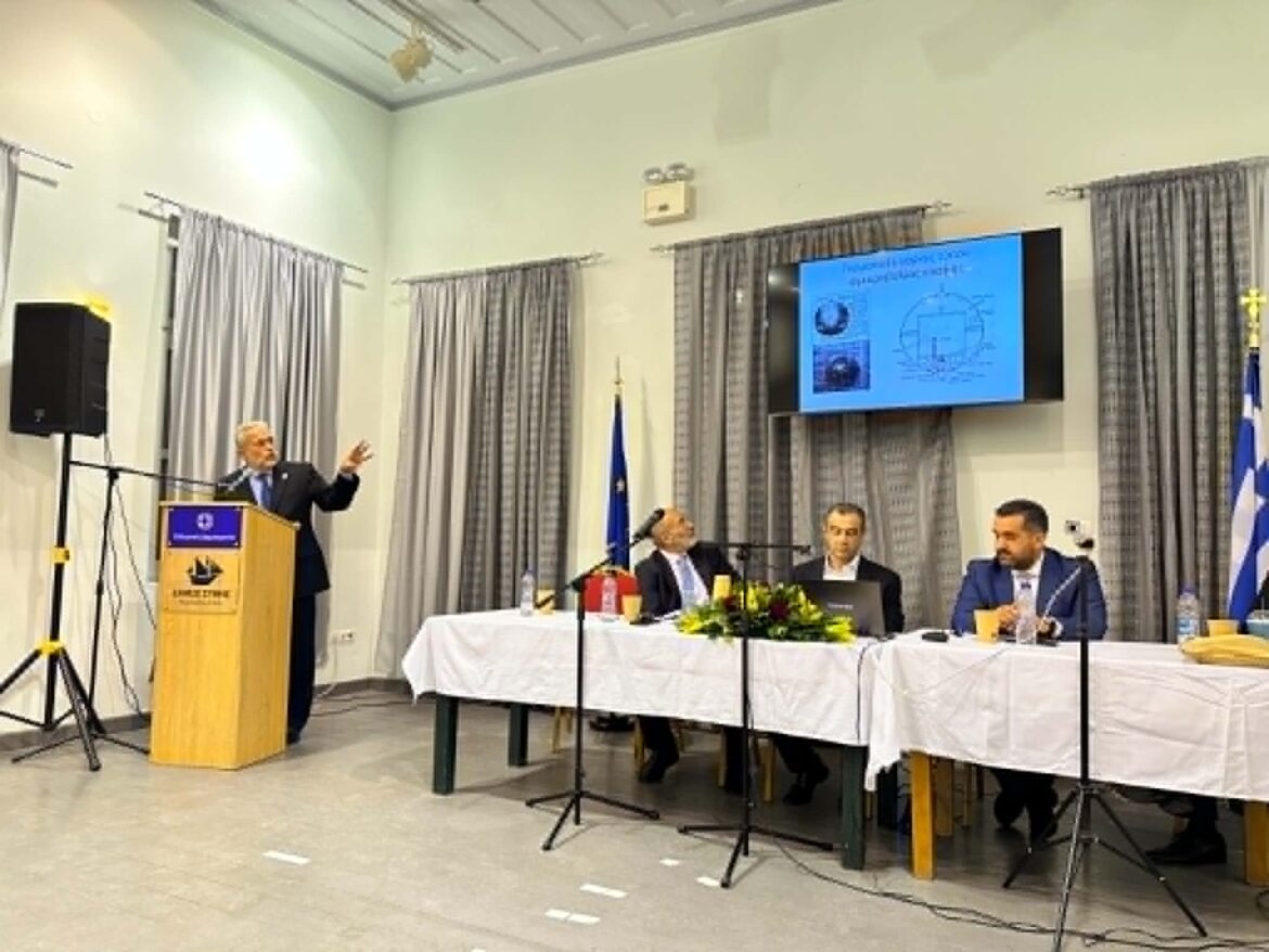 Speaker at a podium addressing a panel of four seated at a long table in a conference room, with a blue presentation screen on the wall behind them.