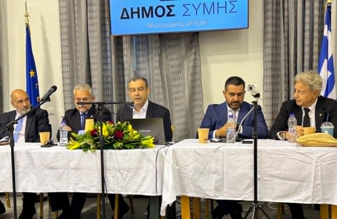 Five men in suits seated at a conference panel with microphones, flowers, and drinks on the table; Greek signage visible above.