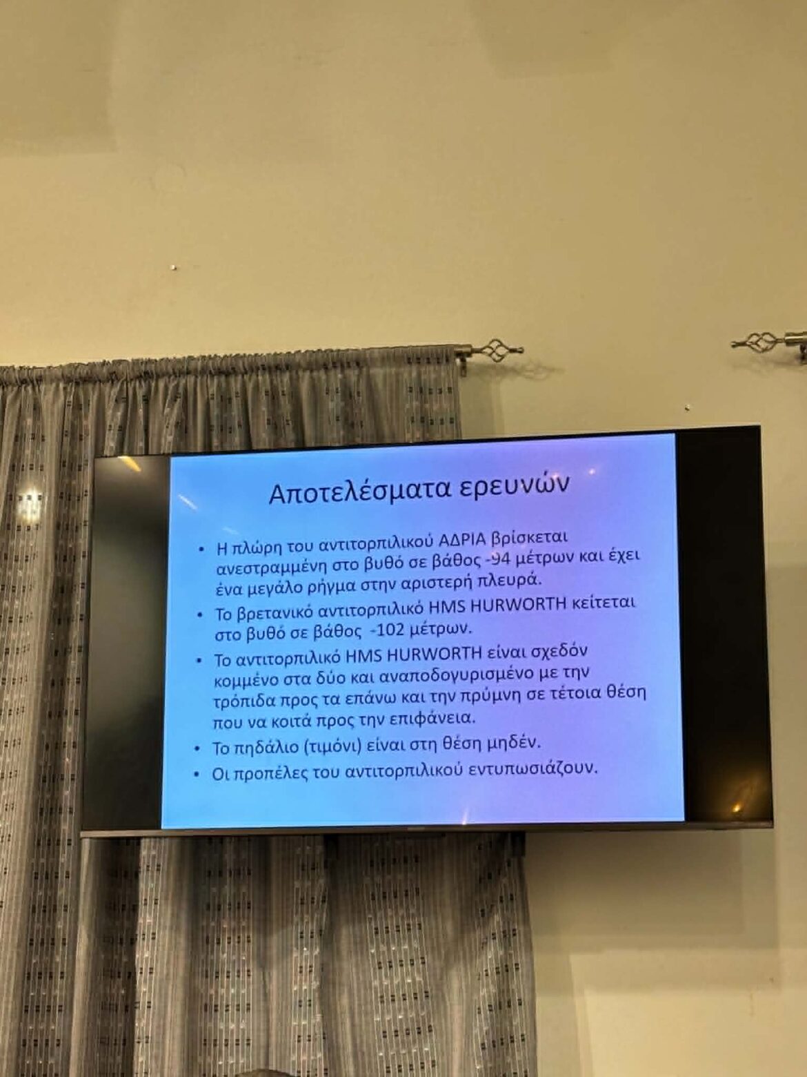 Flat-screen TV on a wall showing a Greek slide titled Αποτελέσματα ερευνών with bullet points, and patterned curtains nearby