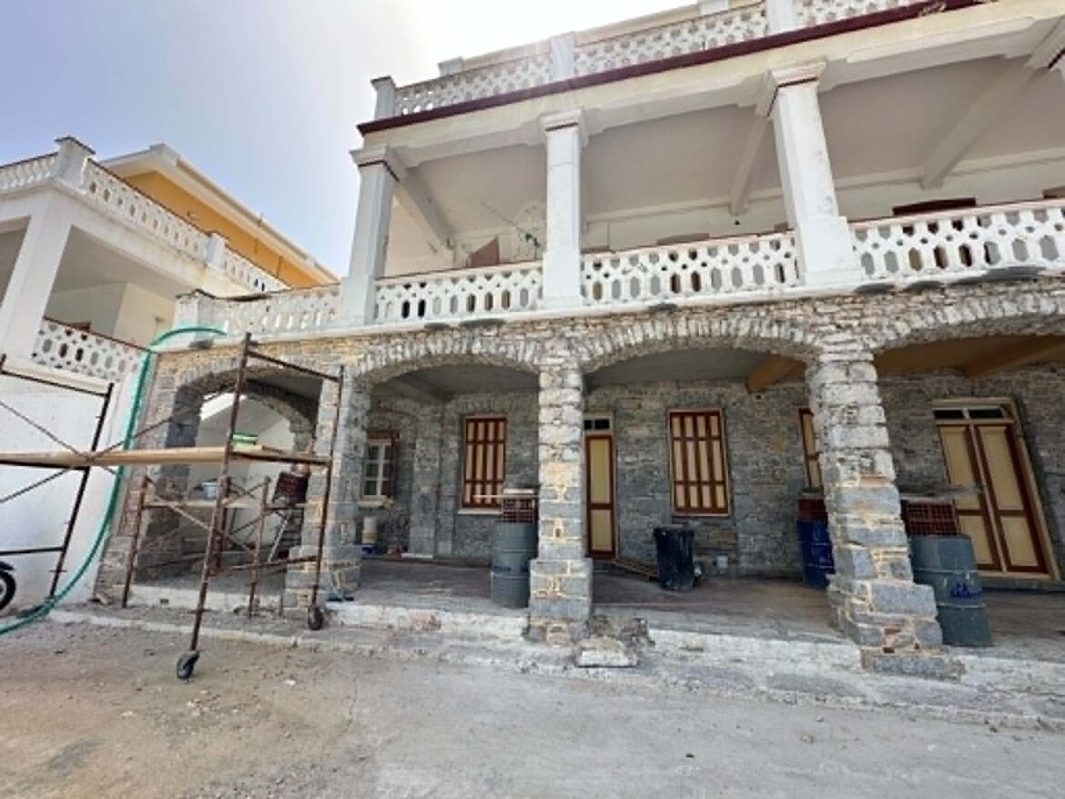 White two-story building with stone columns and decorative balustrades; visible scaffolding and gravel yard in front.
