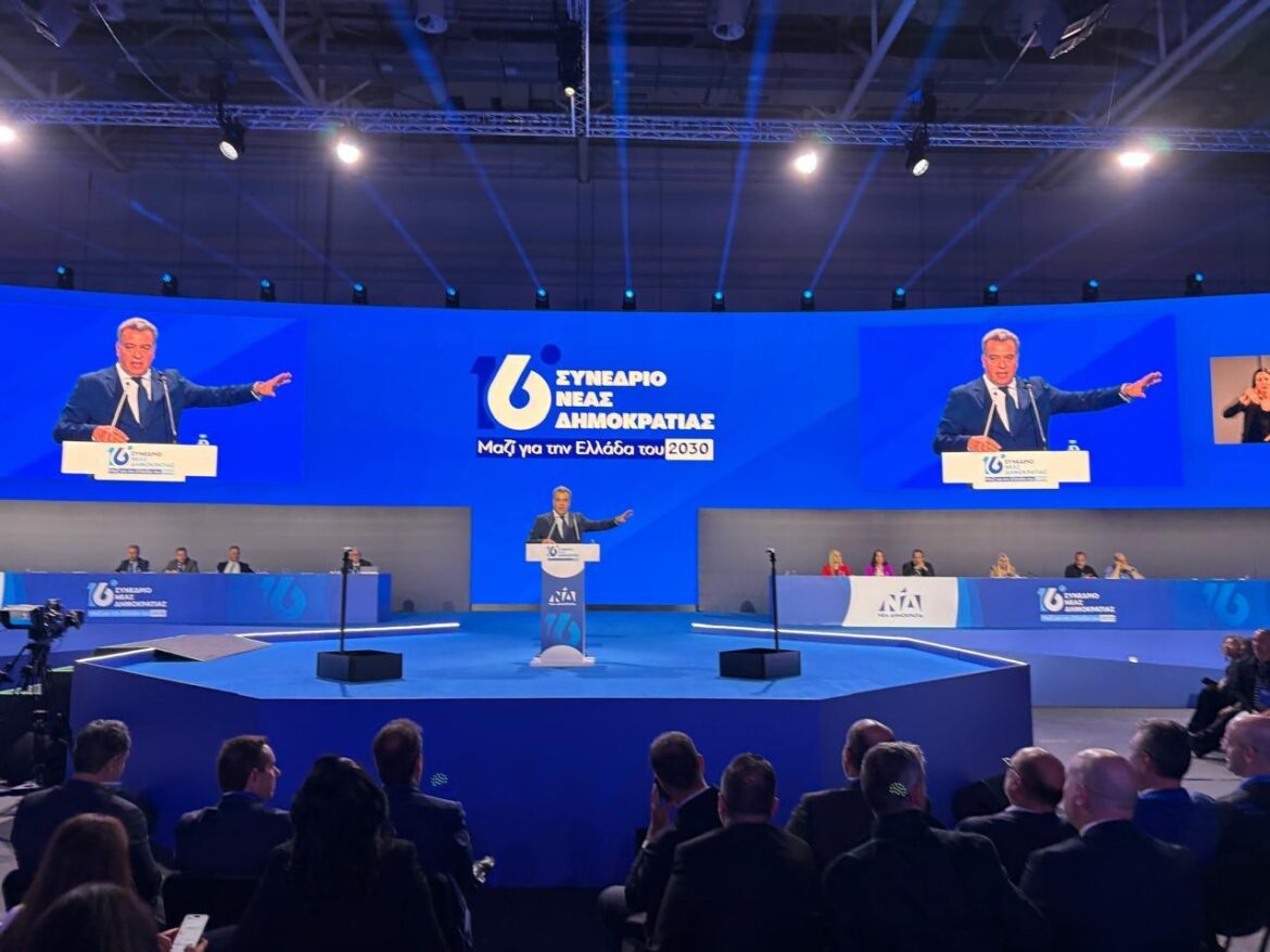 Large blue conference stage with a speaker at a podium, projecting onto three big screens showing the same speaker, Greek text in the center, and an audience seated in front.