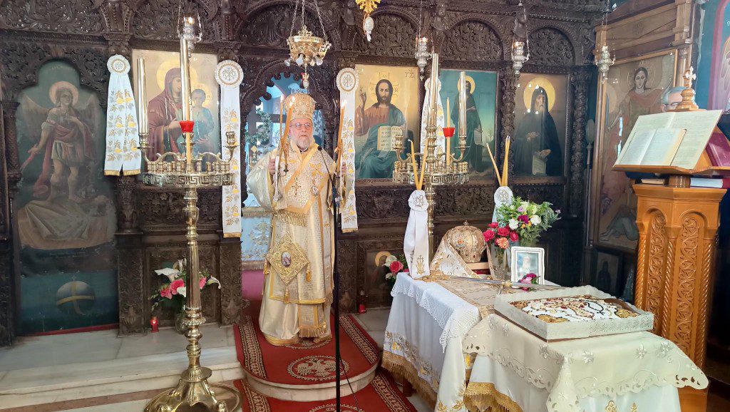 Orthodox priest in gold vestments standing before an iconostasis with lit candles inside an ornate church altar.