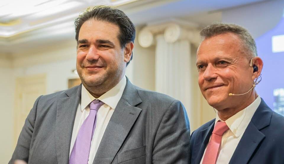 Two men in suits at a conference posing for a photo; one wears a pink tie and a headset mic, both smiling in a bright room with columns in the background
