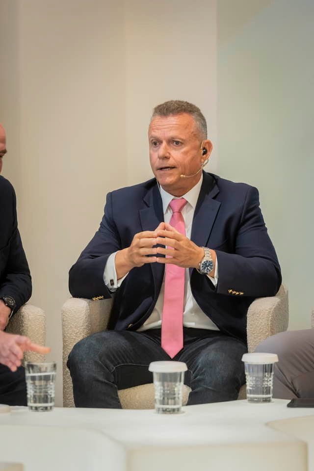 Panelist in a navy suit and pink tie gestures while speaking into a headset microphone.