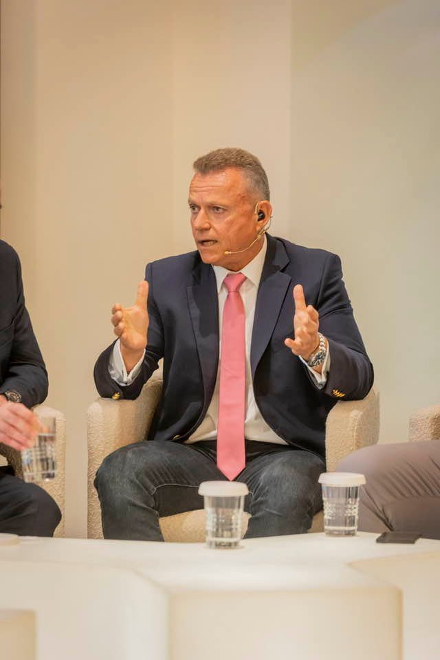 Man in a navy suit and pink tie speaks in a panel, wearing a headset mic and gesturing with his hands.