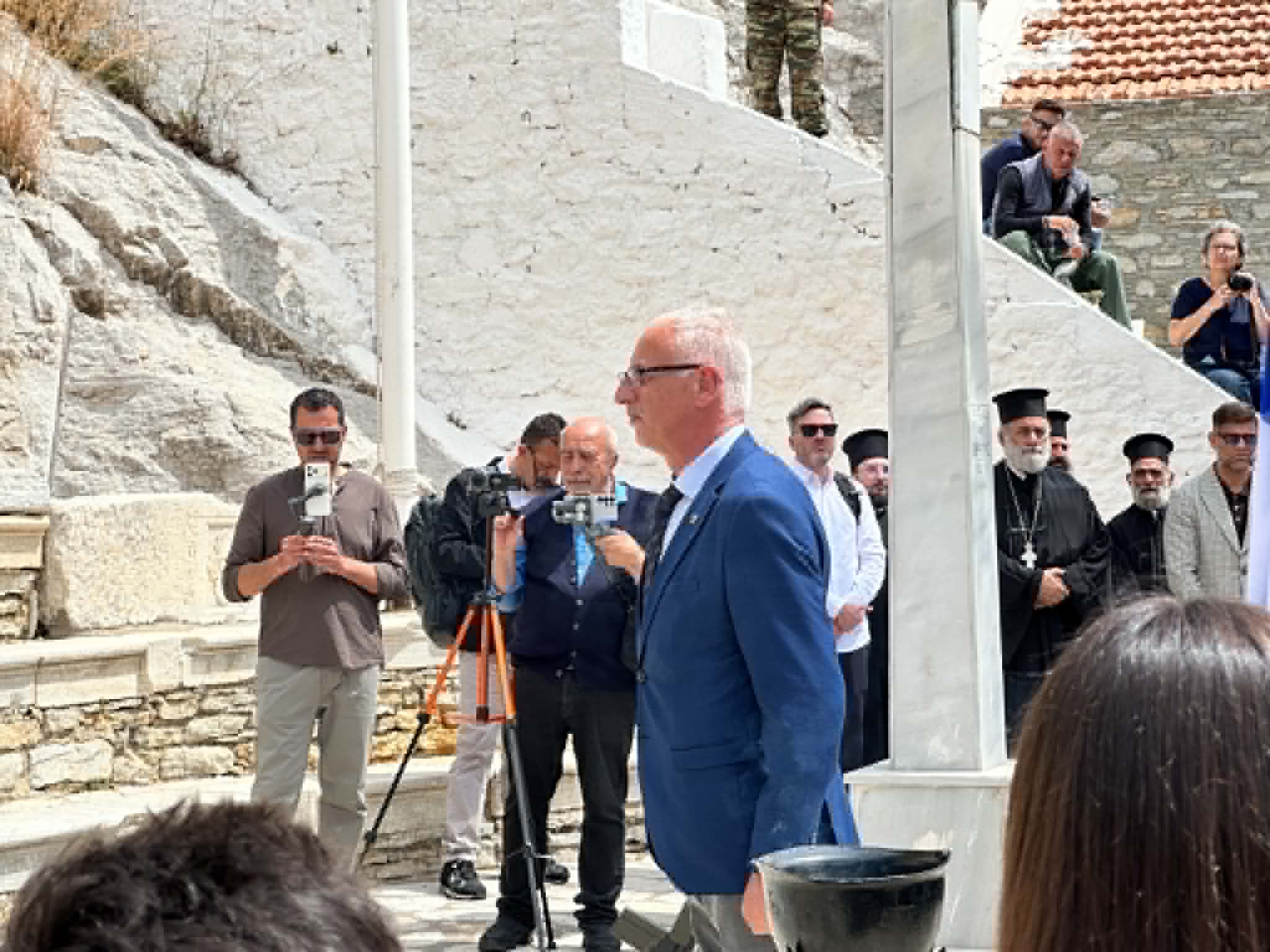 Man in a blue suit speaks into a microphone at an outdoor ceremony, surrounded by reporters and onlookers on stone steps.