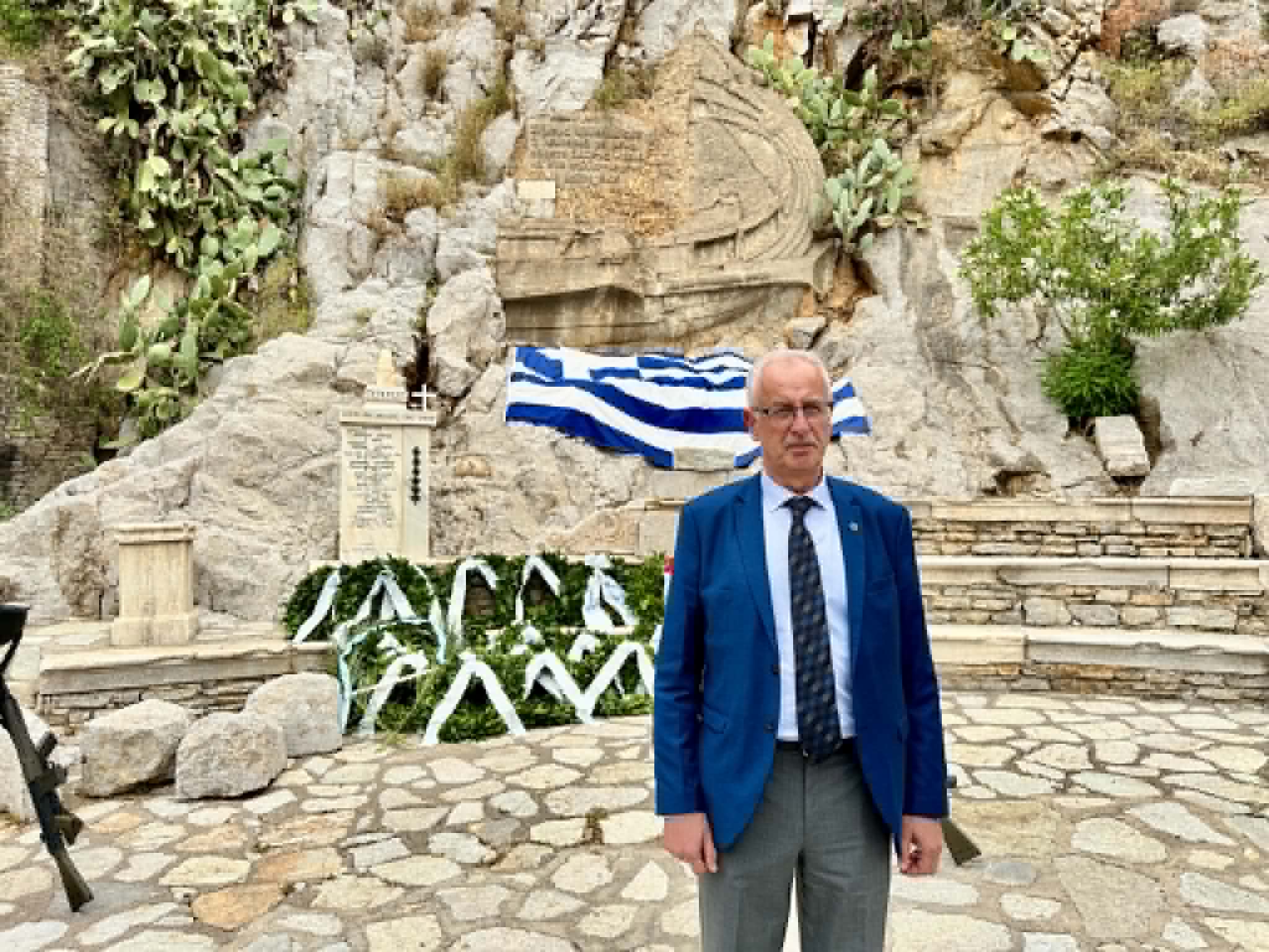 Adult man in a blue suit and patterned tie stands outdoors at a rocky memorial with wreaths behind him.