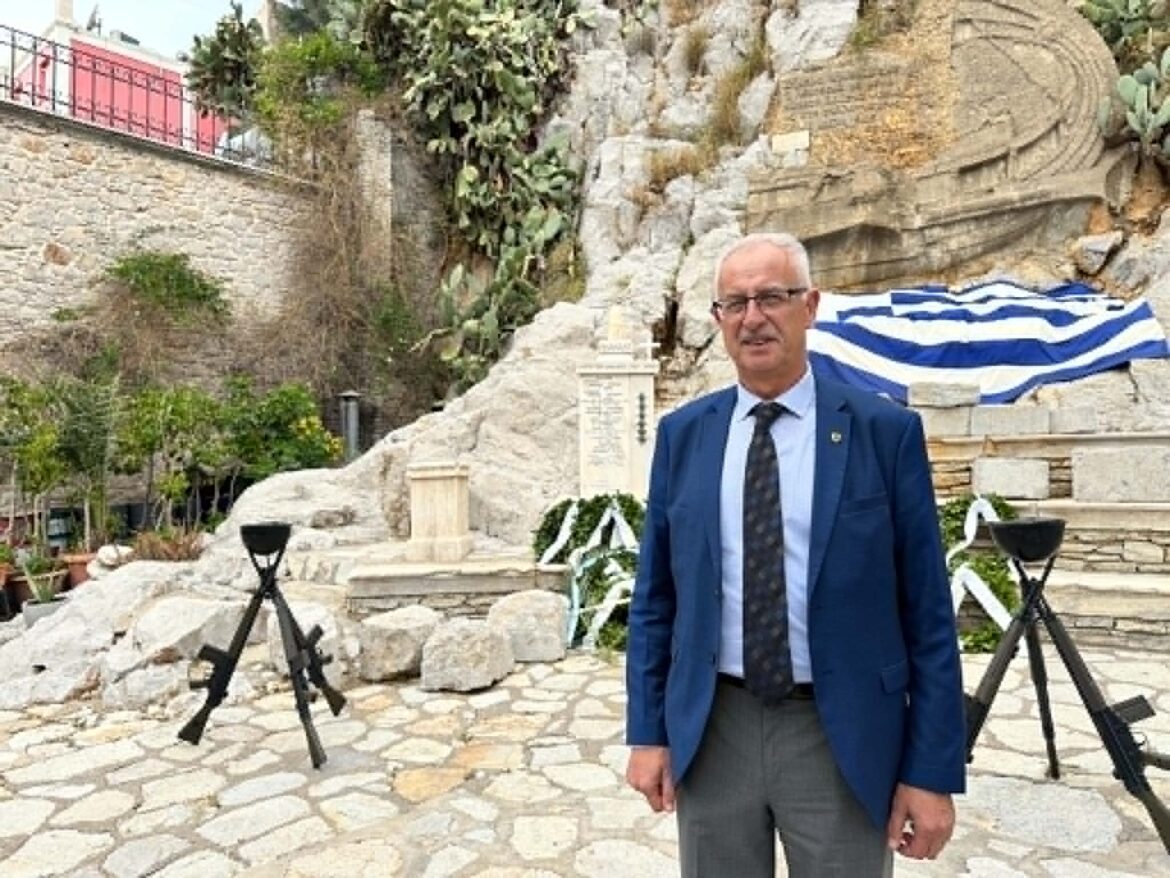 Man in a blue suit standing outdoors in a rocky courtyard with a striped blue-and-white flag in the background, facing the camera.