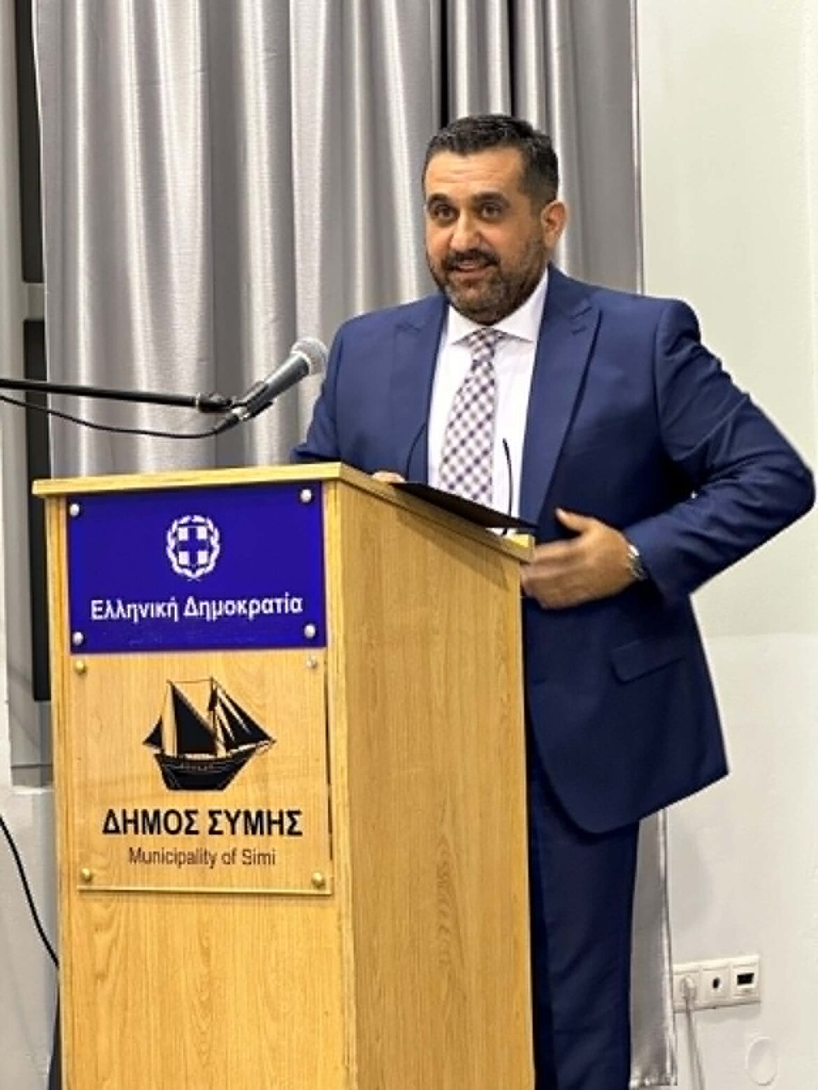 Man in a navy suit giving a talk at a wooden podium with a blue emblem, Greek text and ship symbol visible on the front panel indicating the Municipality of Symi, Greece.