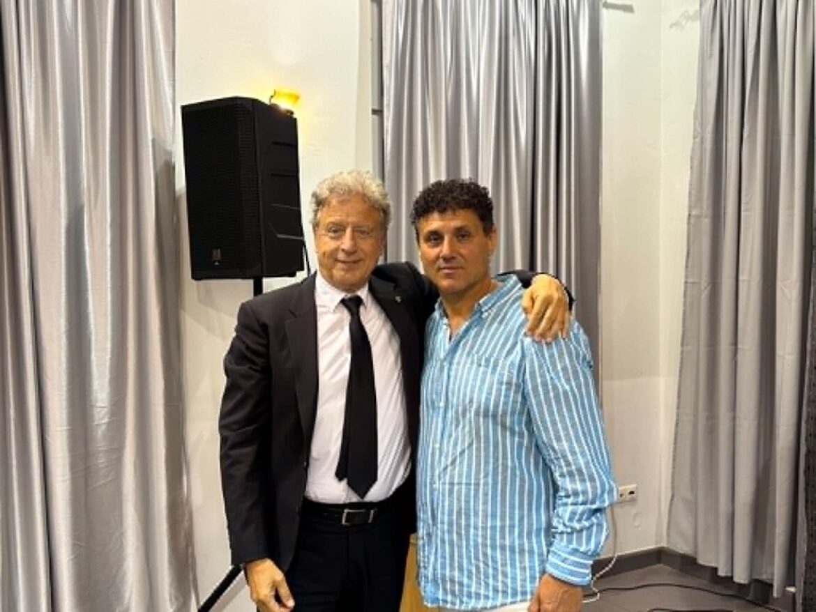 Two men pose together indoors, one in a suit and tie and the other in a blue striped shirt, with a speaker and curtains behind them.