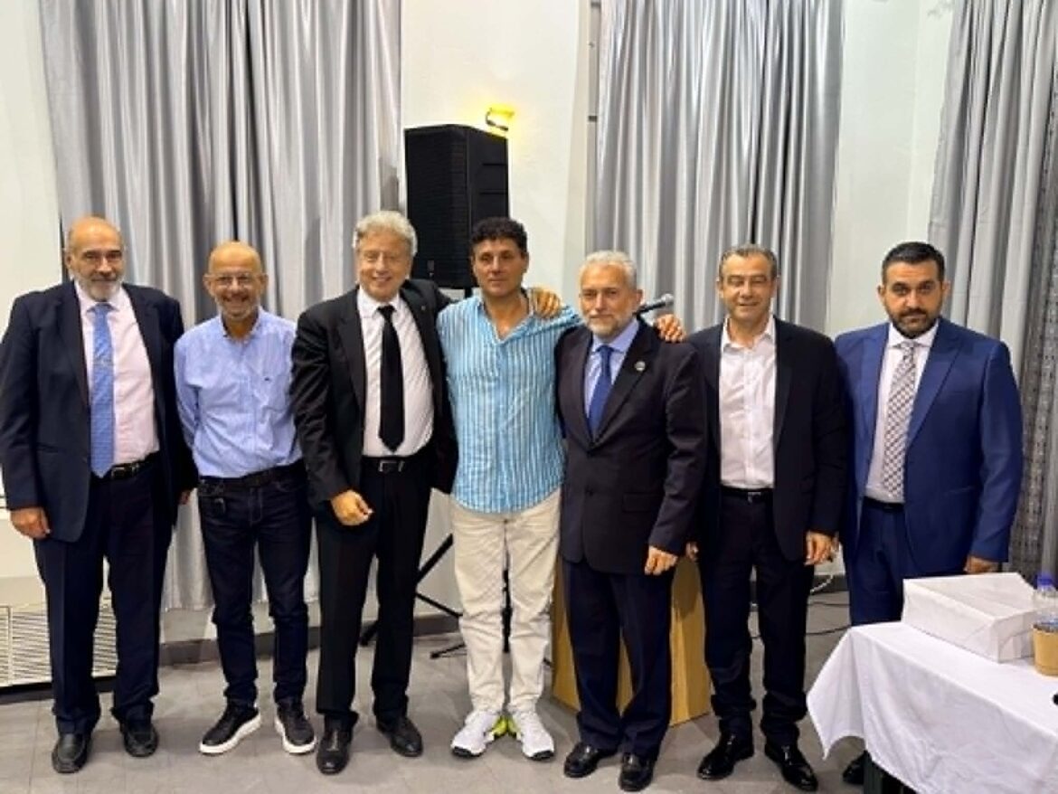 Seven men in business attire standing side by side and posing for a photo in a conference room with grey curtains behind them.