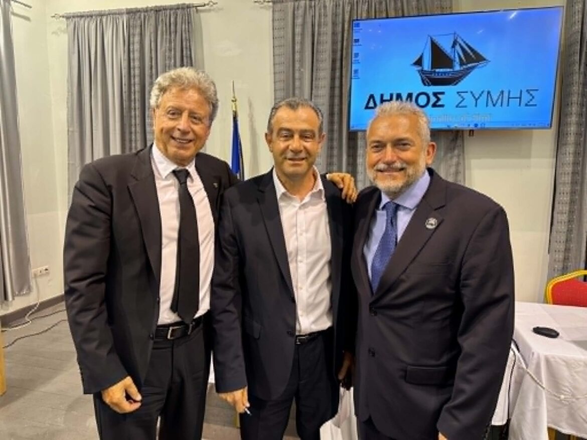 Three men in suits posing together at an indoor event, with a blue screen displaying Greek text in the background.