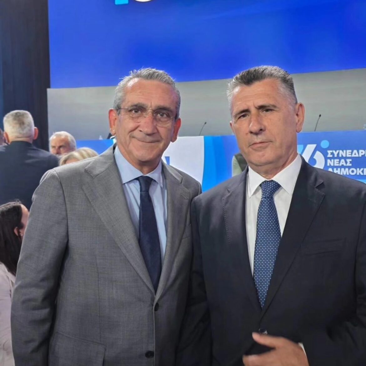 Two men in suits posing for a photo at a formal event, blue stage backdrop behind them.