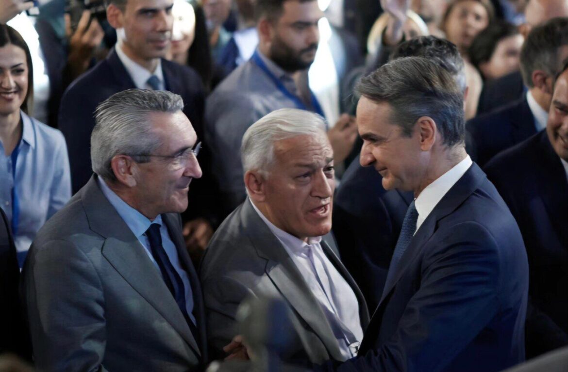Three men in suits shake hands in a crowded event, with onlookers taking photos in the background.