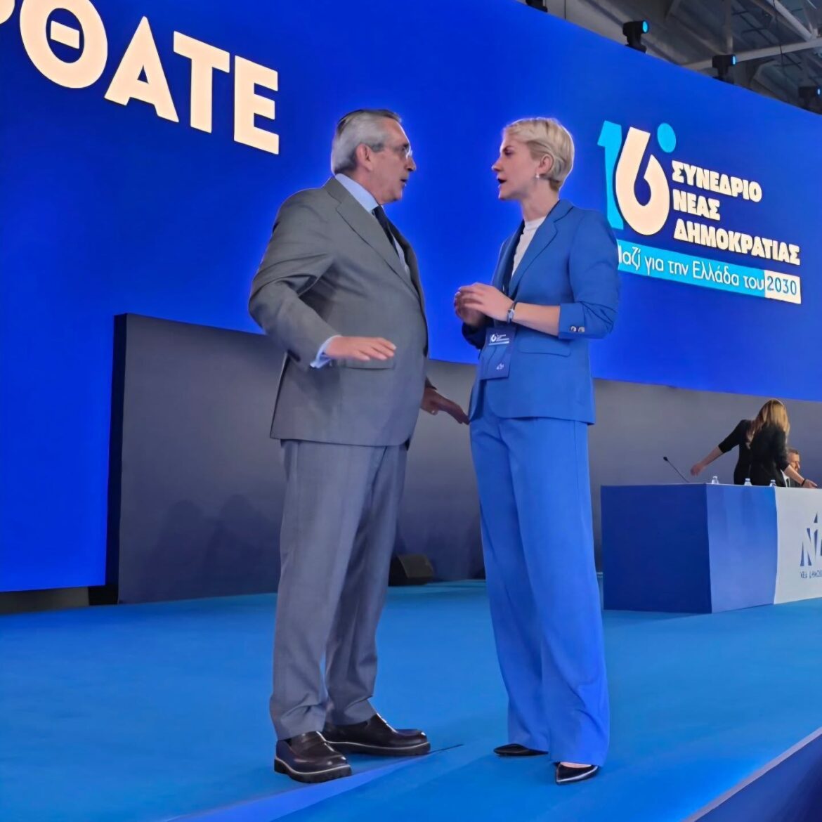 Man in a gray suit and a woman in a blue suit speak to each other on a blue conference stage, with Greek text on the backdrop behind them.
