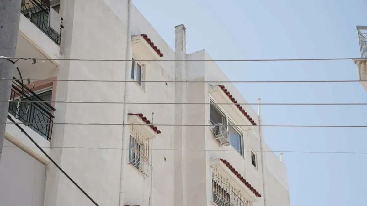 White multi-story residential building with red-tiled accents, barred balconies, and a clear blue sky, wires crossing the scene