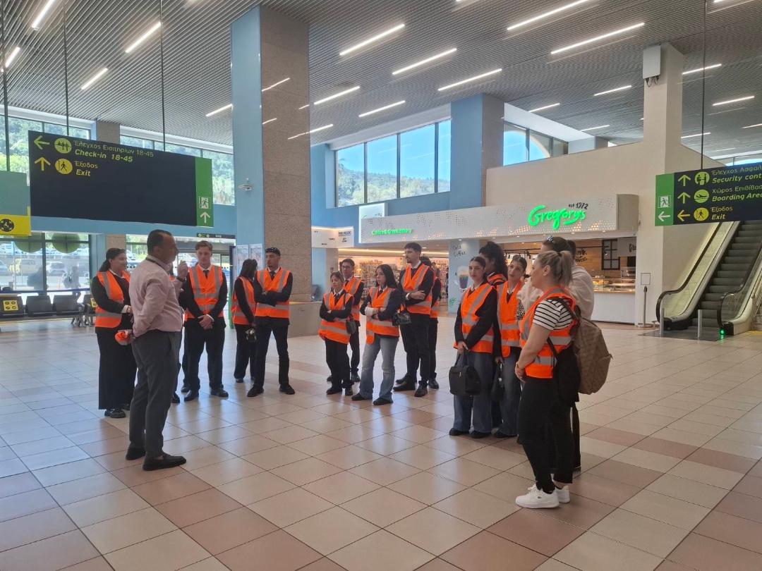 Group of people wearing orange safety vests listening to a man in a pink shirt inside a spacious airport terminal.