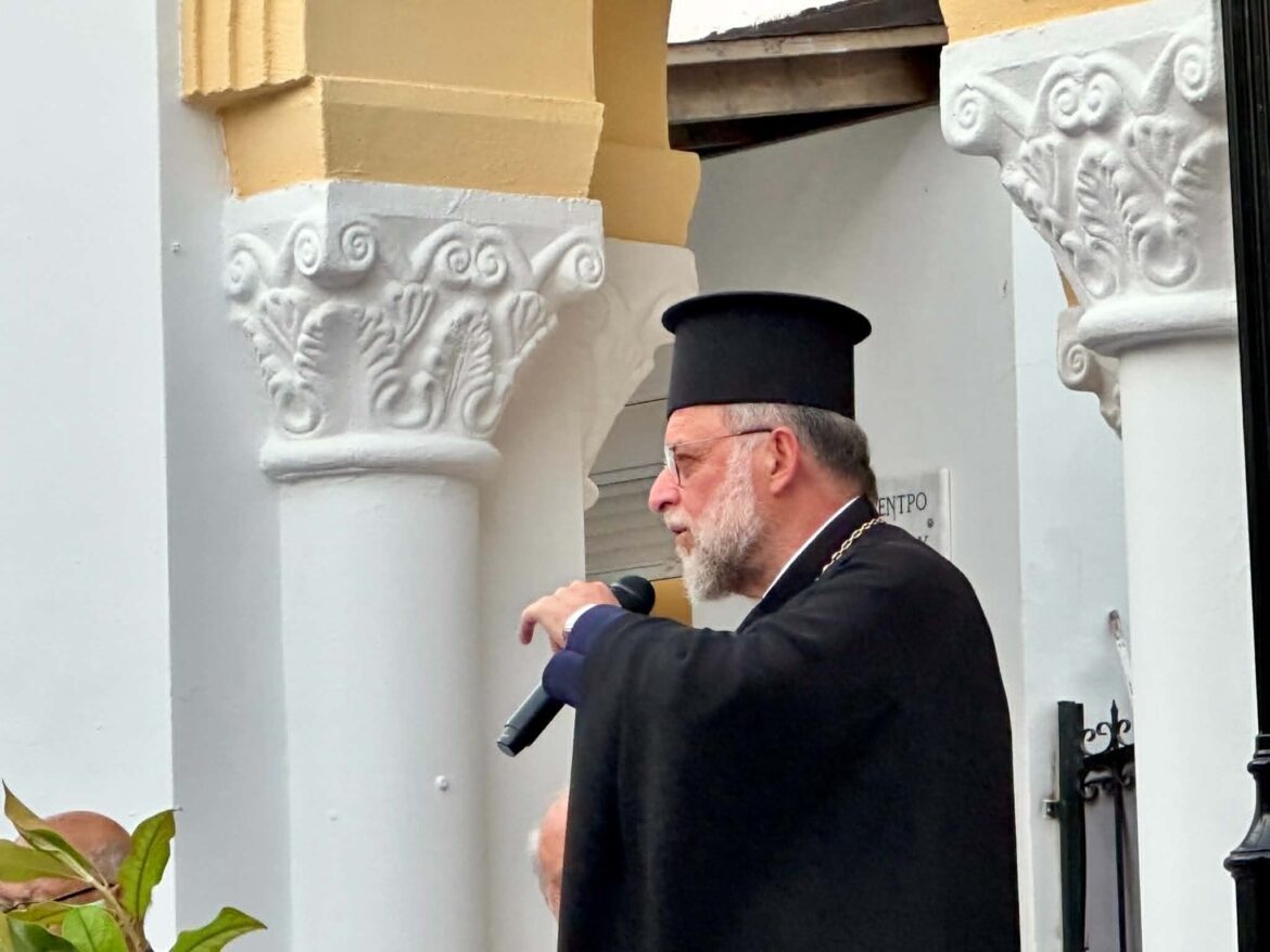 Religious leader in a black suit and tall hat speaking into a handheld microphone beside ornately carved white columns.