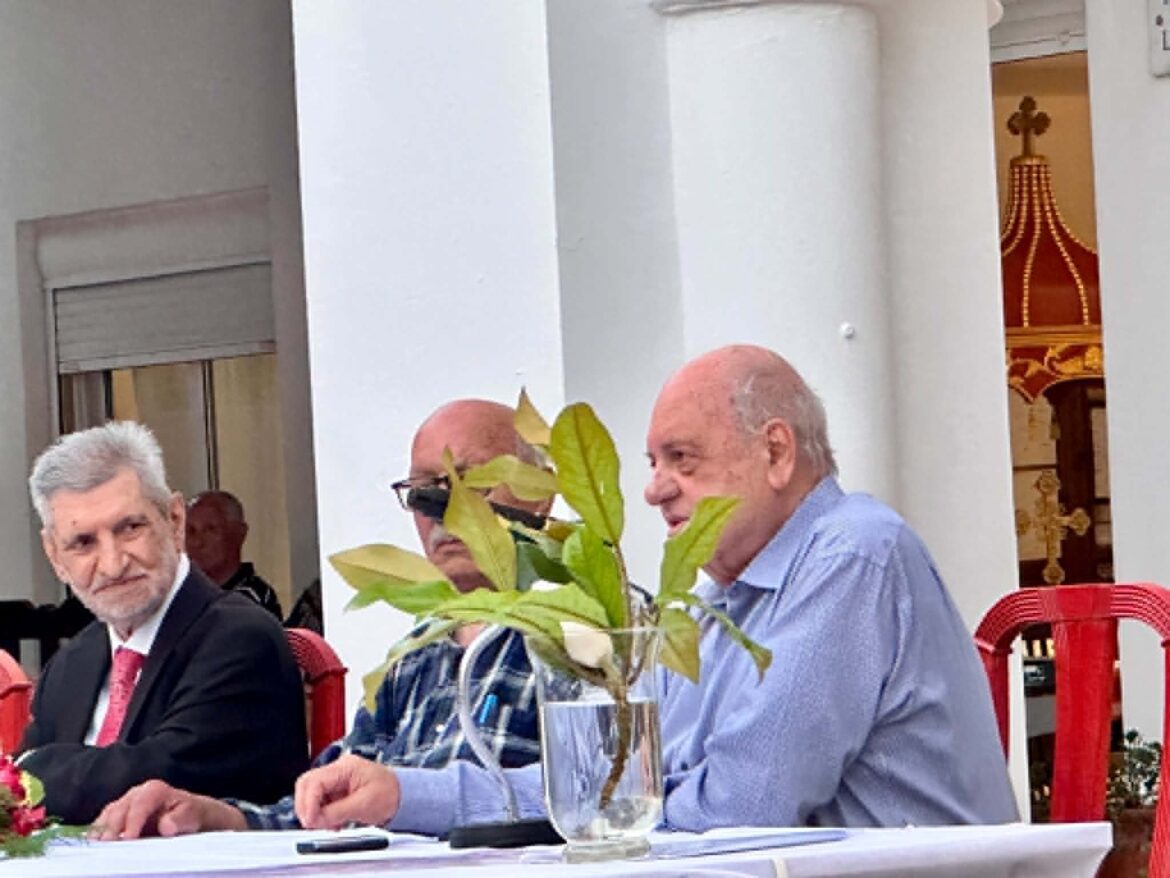 Group of men in formal attire seated at a table, signing documents, with a plant in the foreground.