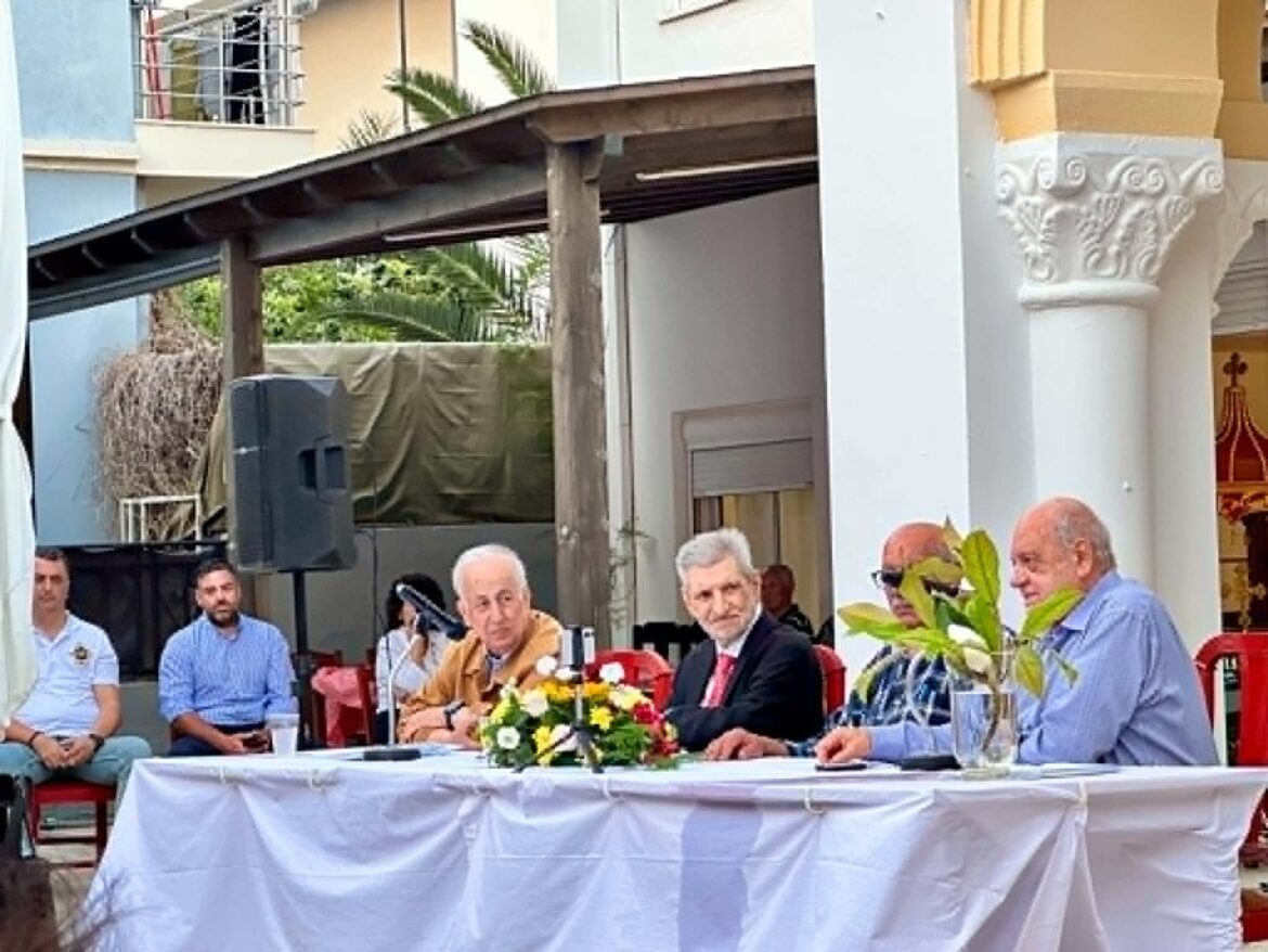 People seated at a long outdoor table for a formal event, with a flower centerpiece in front of them.