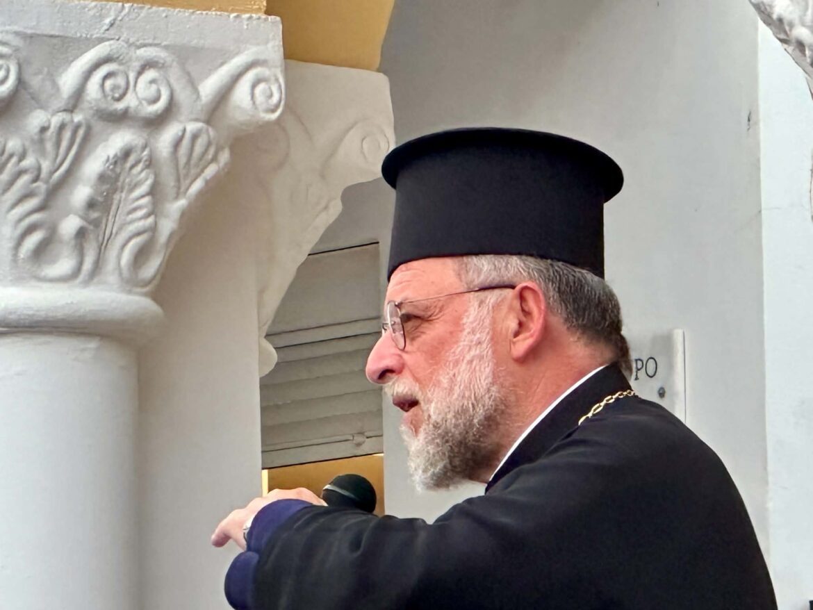 Bearded man in a black clerical hat and glasses stands next to a carved stone column, arms crossed, profile view.