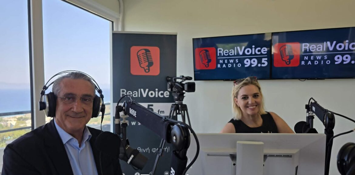 Two radio hosts seated at a broadcast desk with microphones and headphones, in front of RealVoice banners.