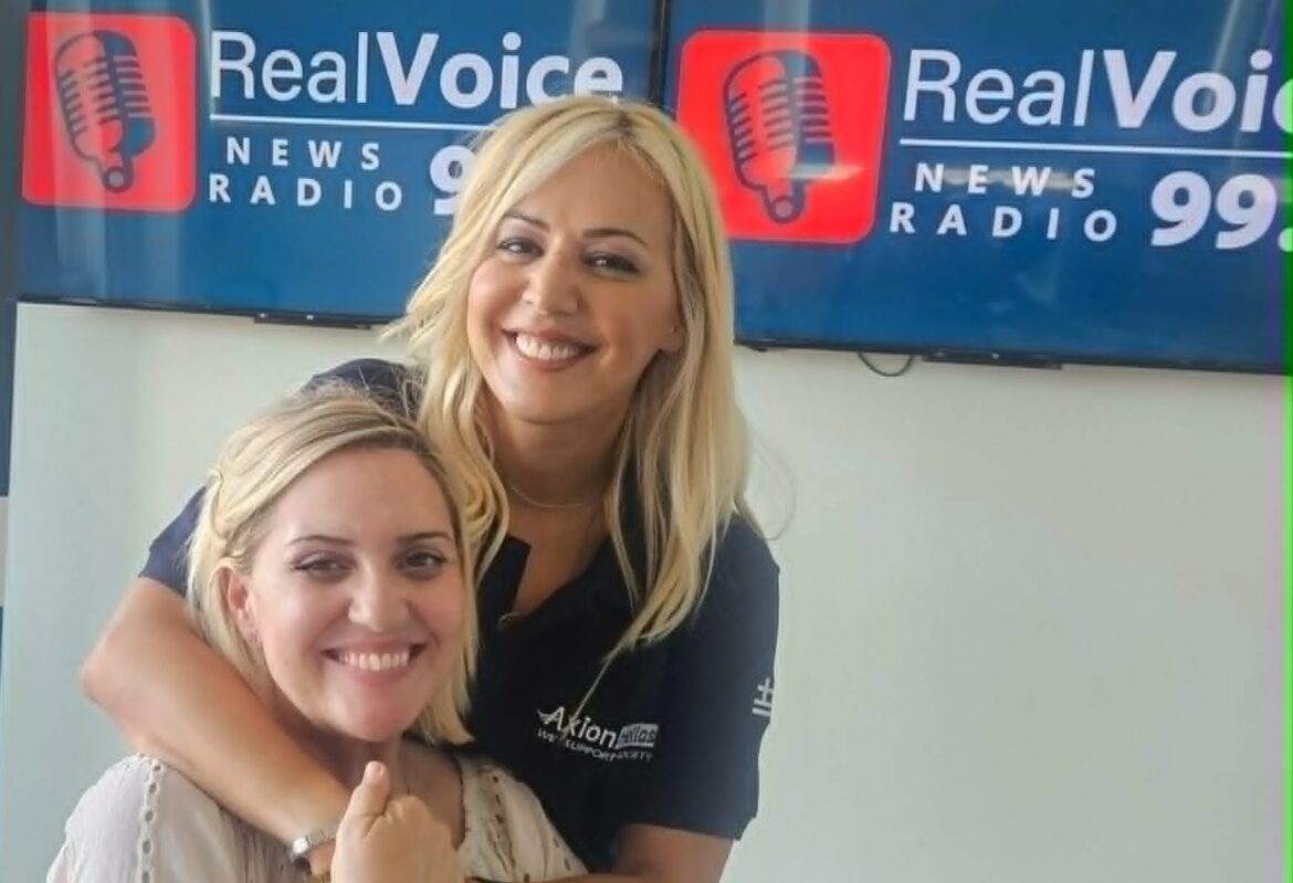 Two women smiling and hugging in a radio studio, with a large microphone in the foreground and a RealVoice backdrop.
