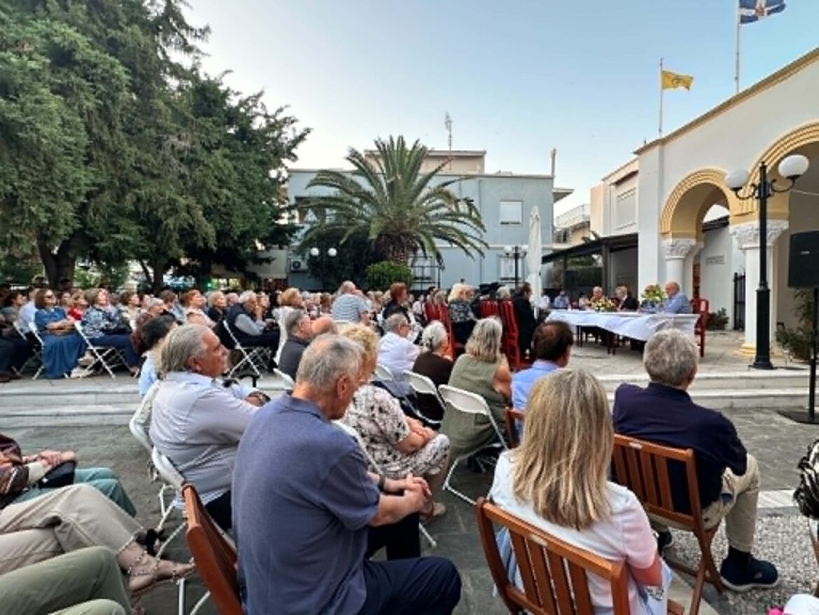 Audience seated in an outdoor plaza for a ceremony, with palm trees and a white-draped table at the front.