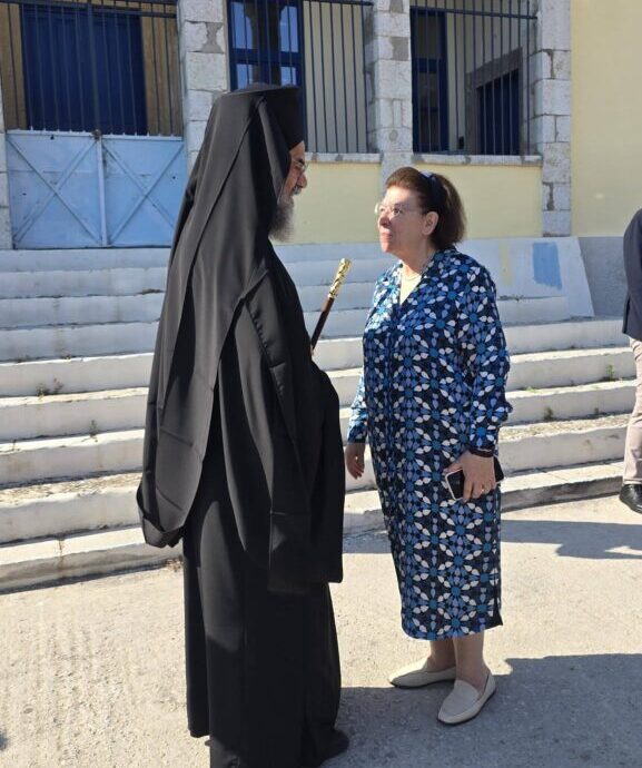 Orthodox nun in a black habit converses with an older woman in a blue floral dress outside a stone building with blue doors and steps.
