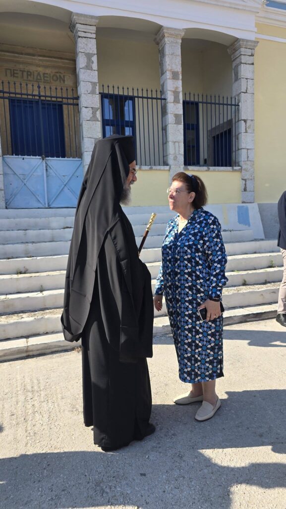 Orthodox nun in a black habit converses with an older woman in a blue floral dress outside a stone building with blue doors and steps.