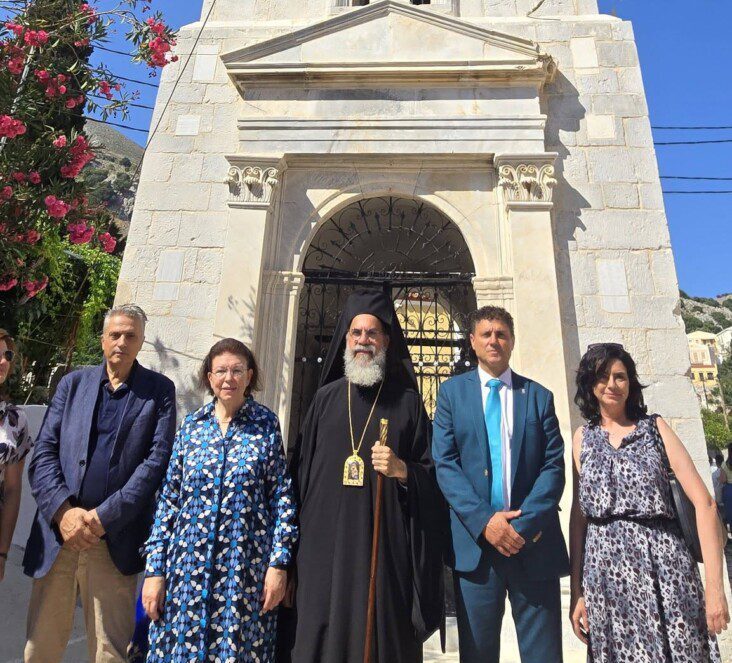 Group of six adults posing in front of a stone church doorway; an Orthodox priest in black robes and cross stands center with a staff, flanked by formally dressed companions.