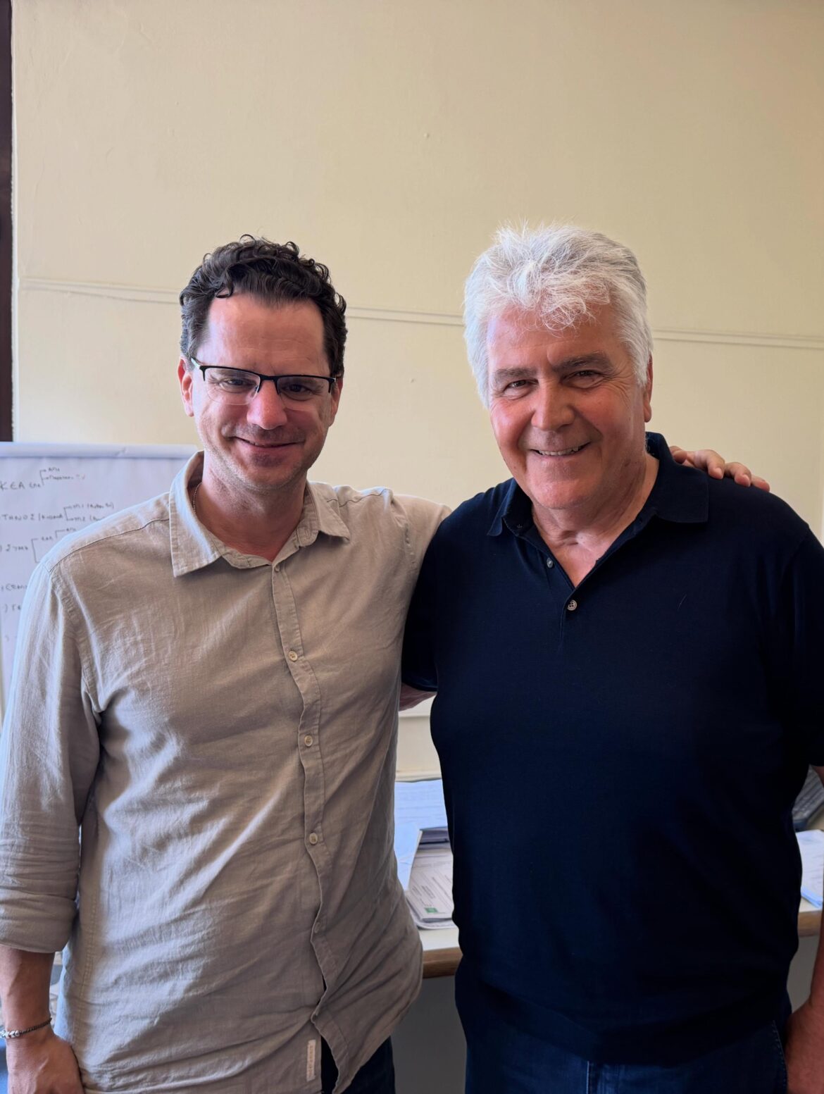 Two men stand indoors with their arms around each other, smiling for the photo.